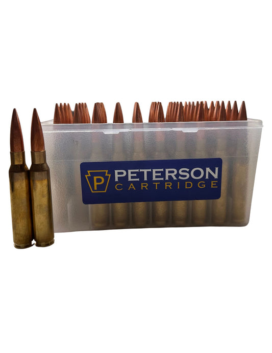 A transparent plastic ammunition box from B&B Firearms containing multiple rounds of .408/419 gr Match rifle ammunition with brass casings and copper nickel alloy bullets. Two additional rounds stand upright next to the box. A blue label on the box reads "Peterson Cartridge".